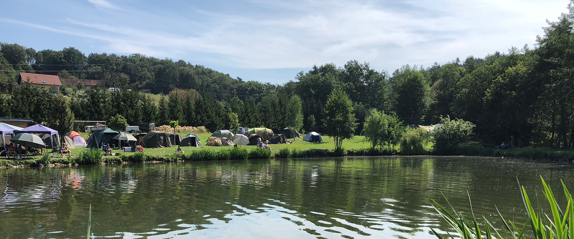 Zeltlager mit mehreren Zelten auf einer Wiese am Ufer eines ruhigen Sees, umgeben von Bäumen und bewaldeten Hügeln im Hintergrund
